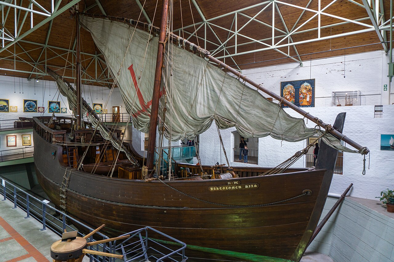 Wooden sailing-ship replica displayed inside a museum, with rigging and a large triangular sail bearing a red cross.
